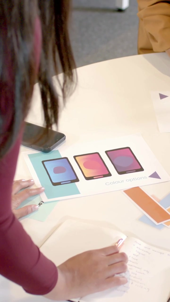 Video verticale: Designer che posiziona un grafico, prende appunti al tavolo e sceglie campioni di colore, indossando una giacca color senape.