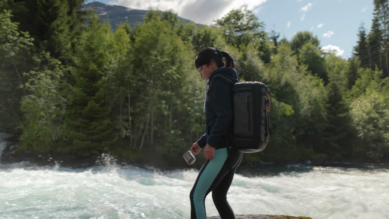 배 ⁇ 을 들고 산 강 근처의 숲에서 자연에서 물을 마시는 여성 여행자.