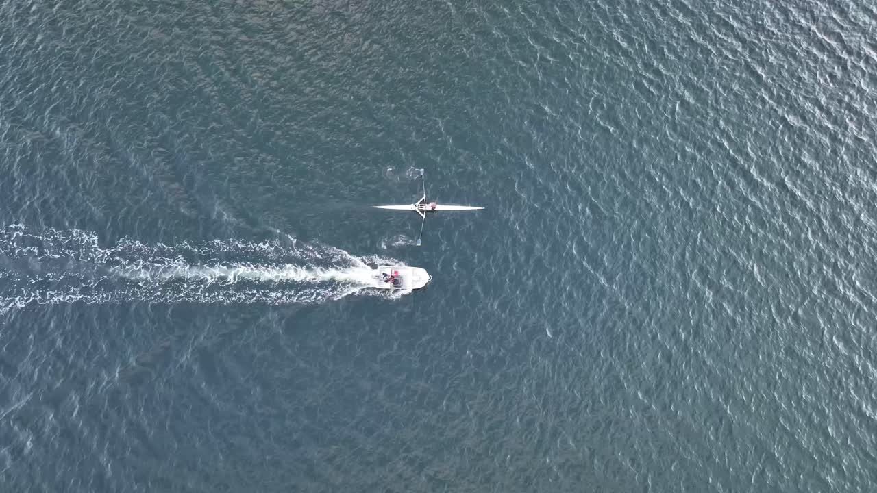 Aerial tracking shot of single rower rowing down a river followed by support boat.