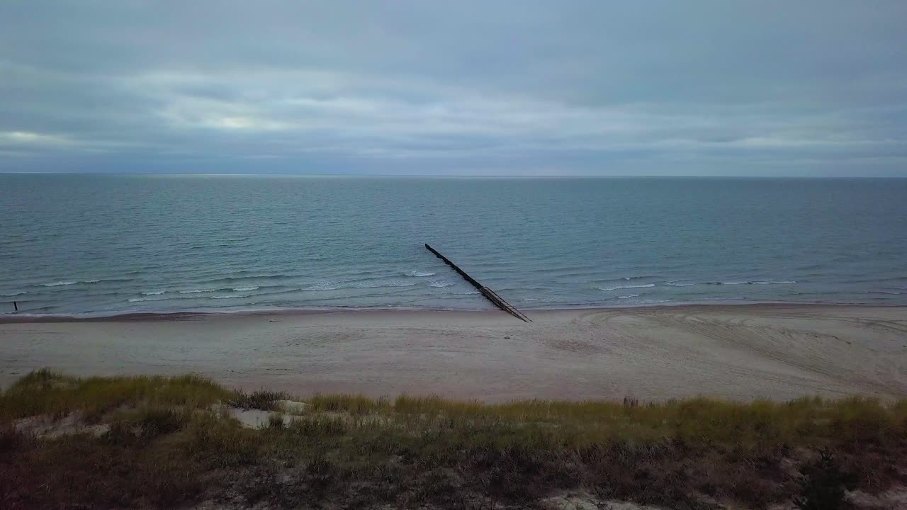 흐린 날 발트해 연안의 공중 전망, 오래된 목조 부두, 하얀 모래 해변, 해안에 부서지는 낮은 파도, 기후 변화, 광각 드론 샷