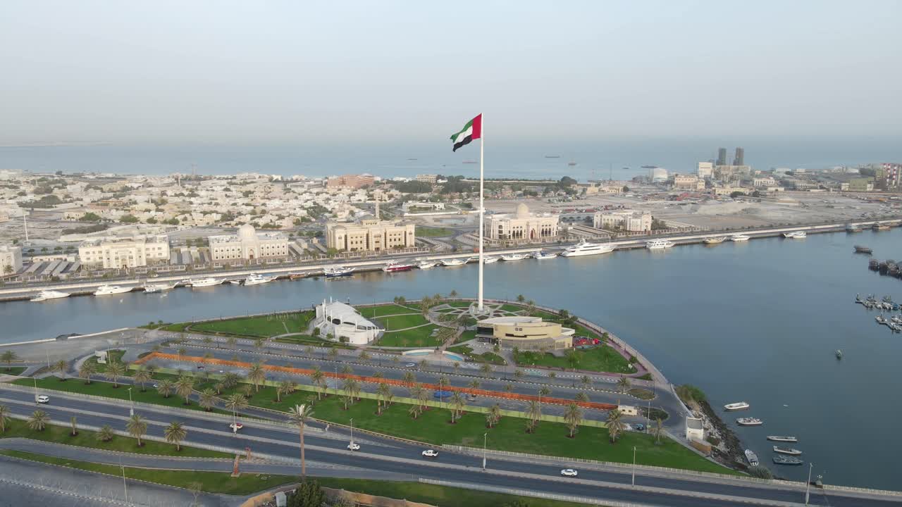 The Flag of the United Arab Emirates waving in the air, the Blue sky and city development in the Background, The national symbol of UAE over Sharjah's Flag Island