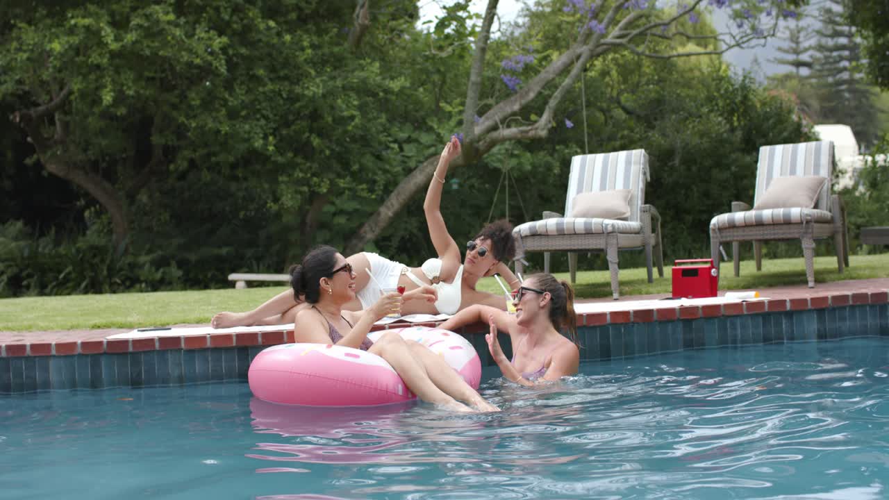 Three women in swimwear passing strawcups on pink float reaching after deck woman raising arm pool