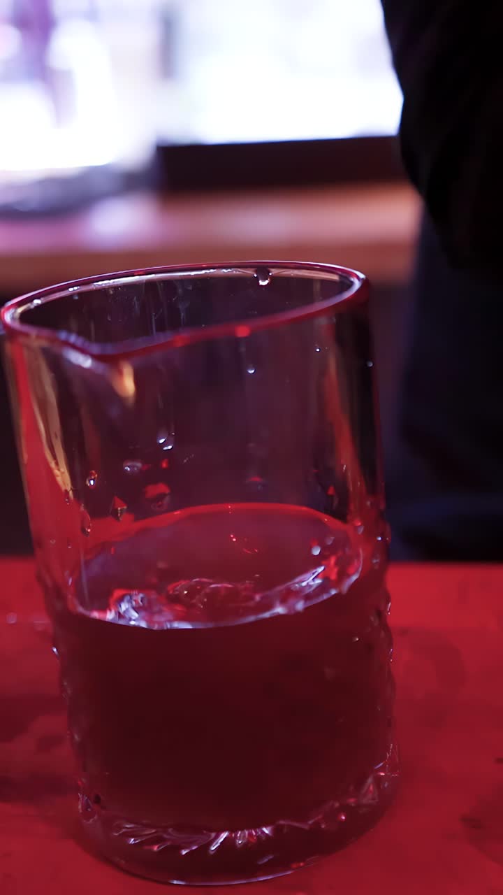 Barman tatuato mescola un cocktail in un bicchiere sotto un'illuminazione rossa, primo piano, telecamera fissa, atmosfera suggestiva