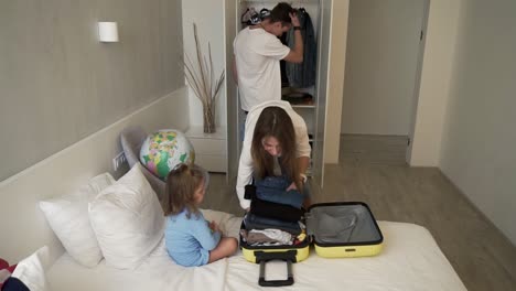 Young-modern-caucasian-couple-gets-ready-for-trip-together-with-their-little-daughter,-packs-suitcase-having-fun,-smiles,-in