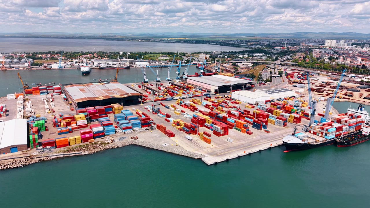 Burgas, Bulgaria, 21 May 2026: Wide angle container port with ships and warehouses, Burgas, Bulgaria. Elevated drone view of vessel at quay, colorful rows and rusty warehouse rooftops.