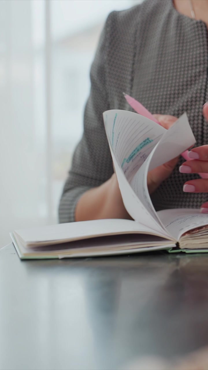 Primo piano di una donna in abbigliamento professionale che sfoglia le pagine di un quaderno, con illuminazione soffusa ed espressione calma. Le sue mani girano il quaderno mentre tiene una penna rosa, rivelando carta a righe