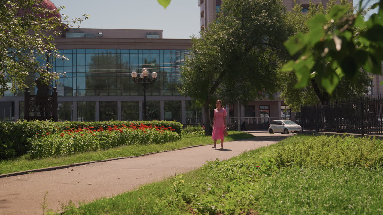 Donna in abito rosa che percorre un sentiero attraverso un tranquillo giardino urbano, edificio in vetro sullo sfondo, aiuole di tulipani che costeggiano il vialetto, luce screziata tra i rami frondosi, atmosfera contemplativa con ritmo cinematografico lento