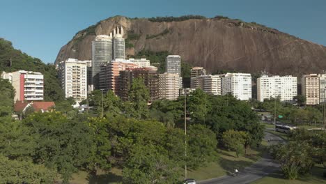 slow aerial ascent with residential buildings and a rock mountain behind revealing a highway through a green environment in rio de janeiro at sunset