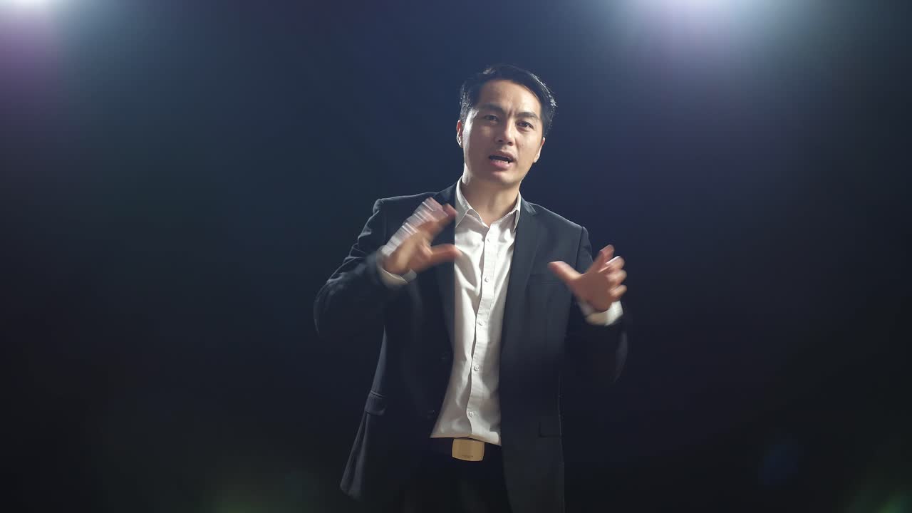 hombre orador asiático en traje de negocios sosteniendo sus manos juntas y mostrando los dedos índice mientras habla en el estudio de pantalla negra