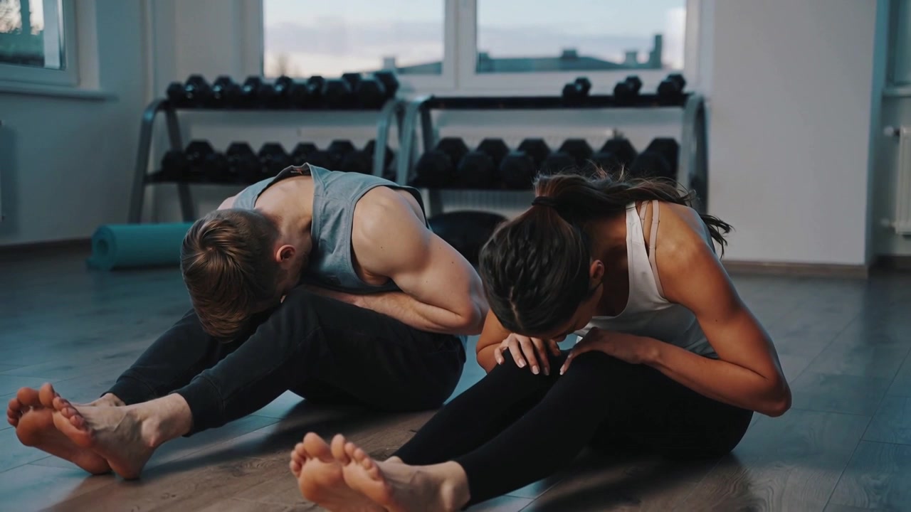 Breathing, pair hinging forward and holding for stretch on mats in studio wearing gray tank