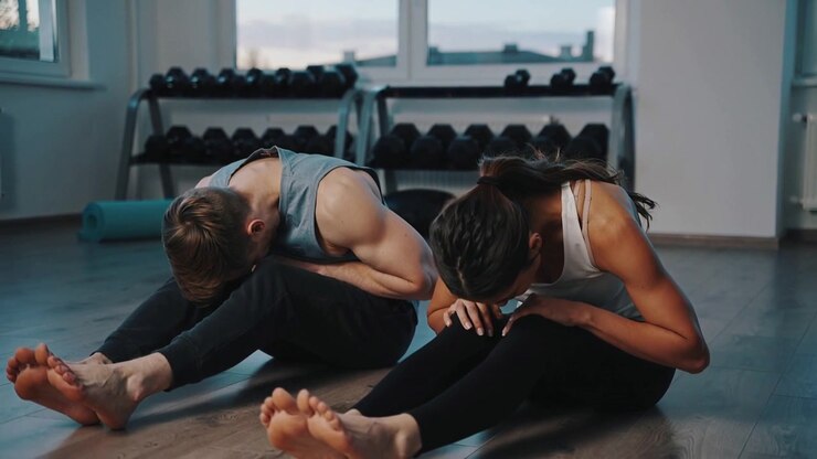 Breathing, pair hinging forward and holding for stretch on mats in studio wearing gray tank