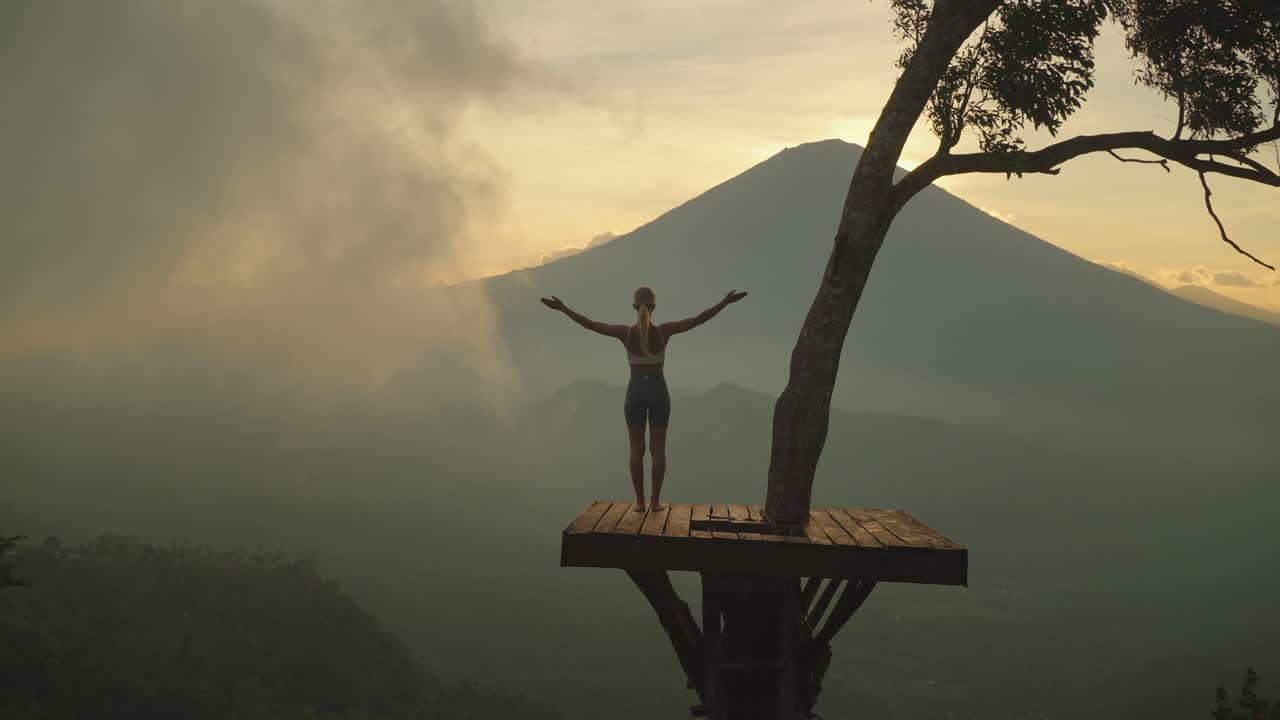 mujer esbelta en una plataforma de árbol de madera levantando los brazos en pose de saludo, monte agung
