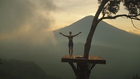 schlanke frau auf hölzerner baumplattform, die arme in begrüßungspose hebt, mount agung