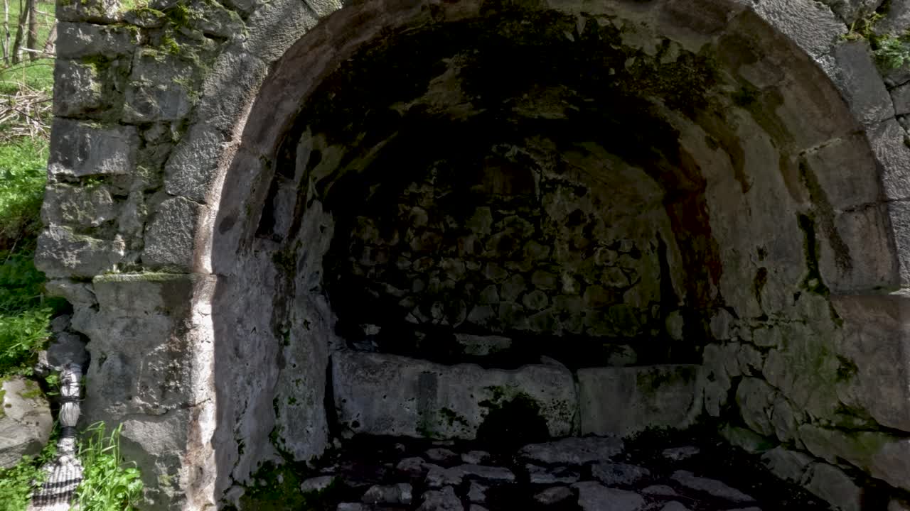 Uma antiga estrutura de pedra fica em uma floresta. A luz do sol brilha através das árvores, destacando o musgo nas pedras. A área parece intocada e tranquila. A natureza cerca este local histórico