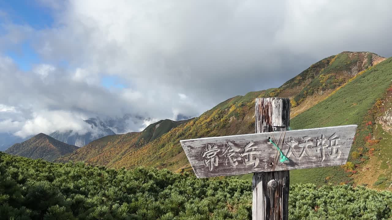 Scatto di un cartello rustico sullo sfondo delle spettacolari Alpi giapponesi settentrionali in autunno, che mette in mostra un fogliame dai colori vivaci. L'ampio angolo rivela un paesaggio montano tranquillo e vasto.