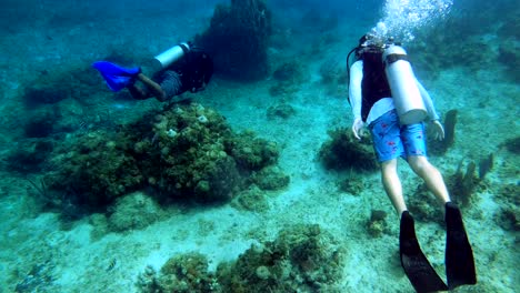 two-scuba-divers-exploring-ocean-floor-coral-reef,-fish-swimming-around,-sand,-rocks,-oxygen-tank-making-bubbles,-wide-shot,-deep-sea-diving,-cinematic