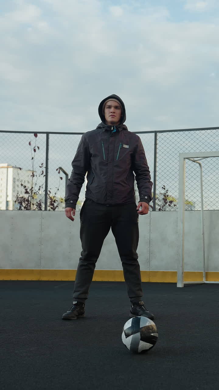 entrenador realizando sentadillas con las manos juntas en preparación para el ejercicio en un campo deportivo al aire libre, con la pelota de fútbol en primer plano, rodeado de edificios residenciales urbanos de gran altura y postes de gol