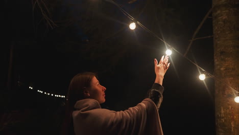 Woman pushes garland of lights in glamping against forest. Young wife enjoys evening tranquility among illuminated forest late at night. Exploring camp vicinity