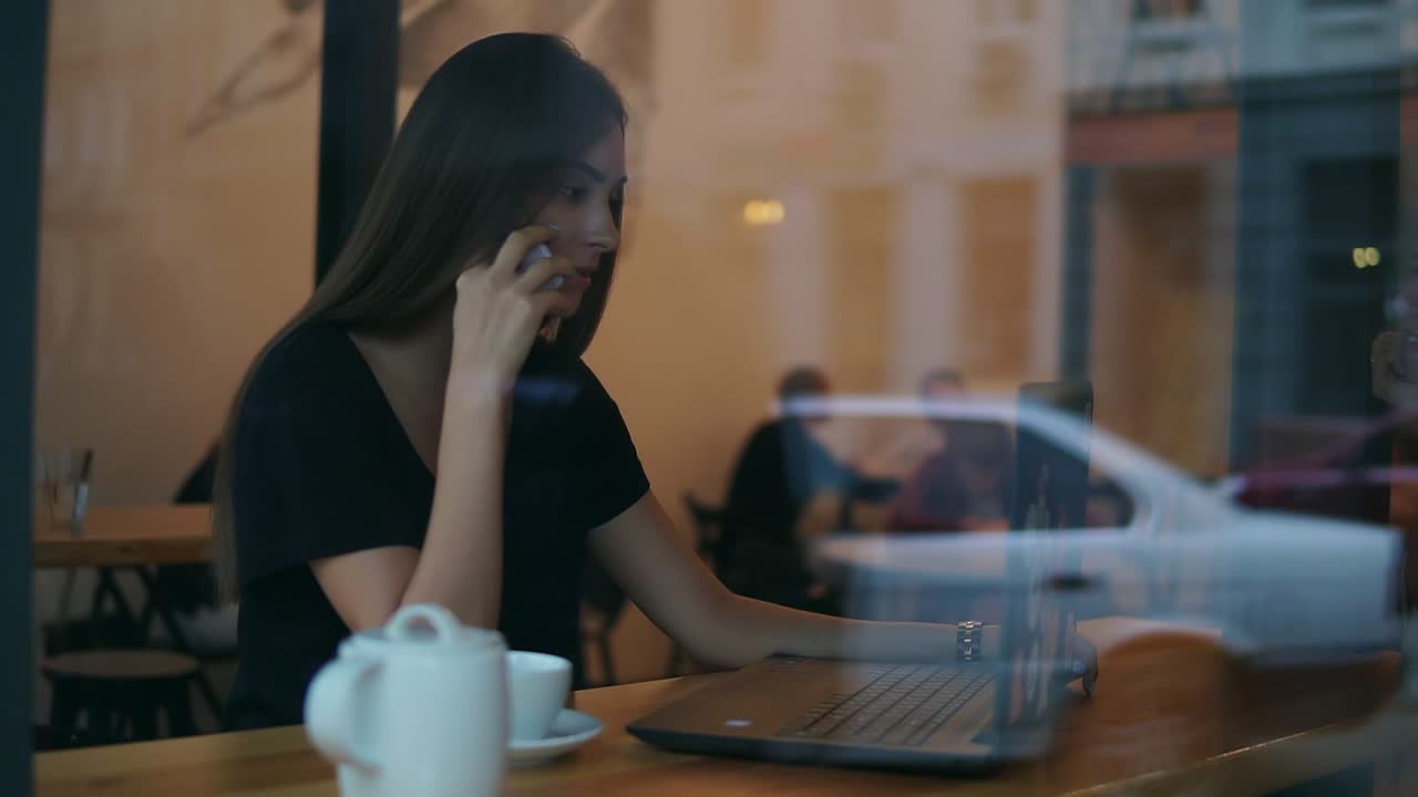 View from the outside of and attractive young woman talking on the phone in a cafe and looking at the screen of her laptop during