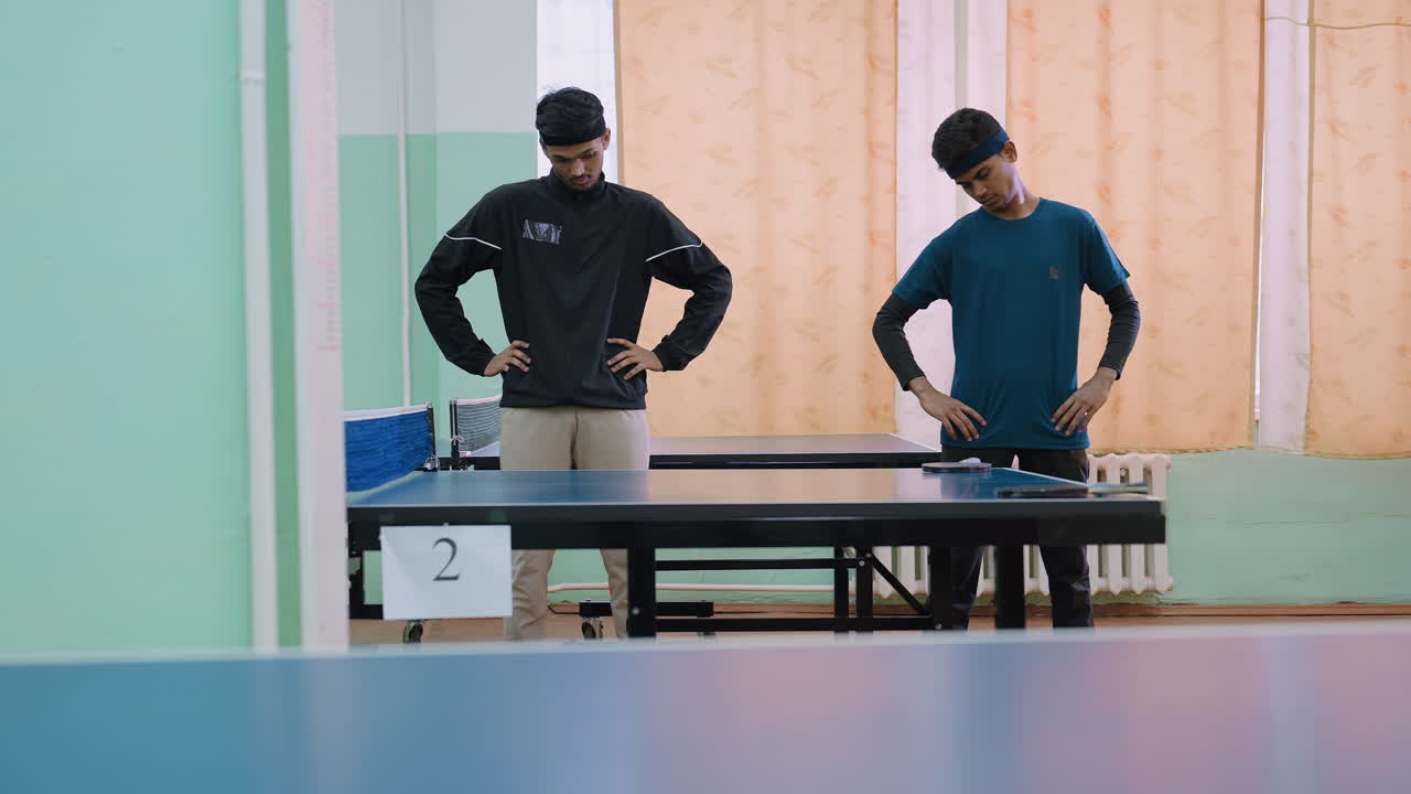 Two young athletes stand beside tennis table performing neck flexibility training, hands on hips, preparing body with warm up session before workout in indoor sports hall with rackets on table