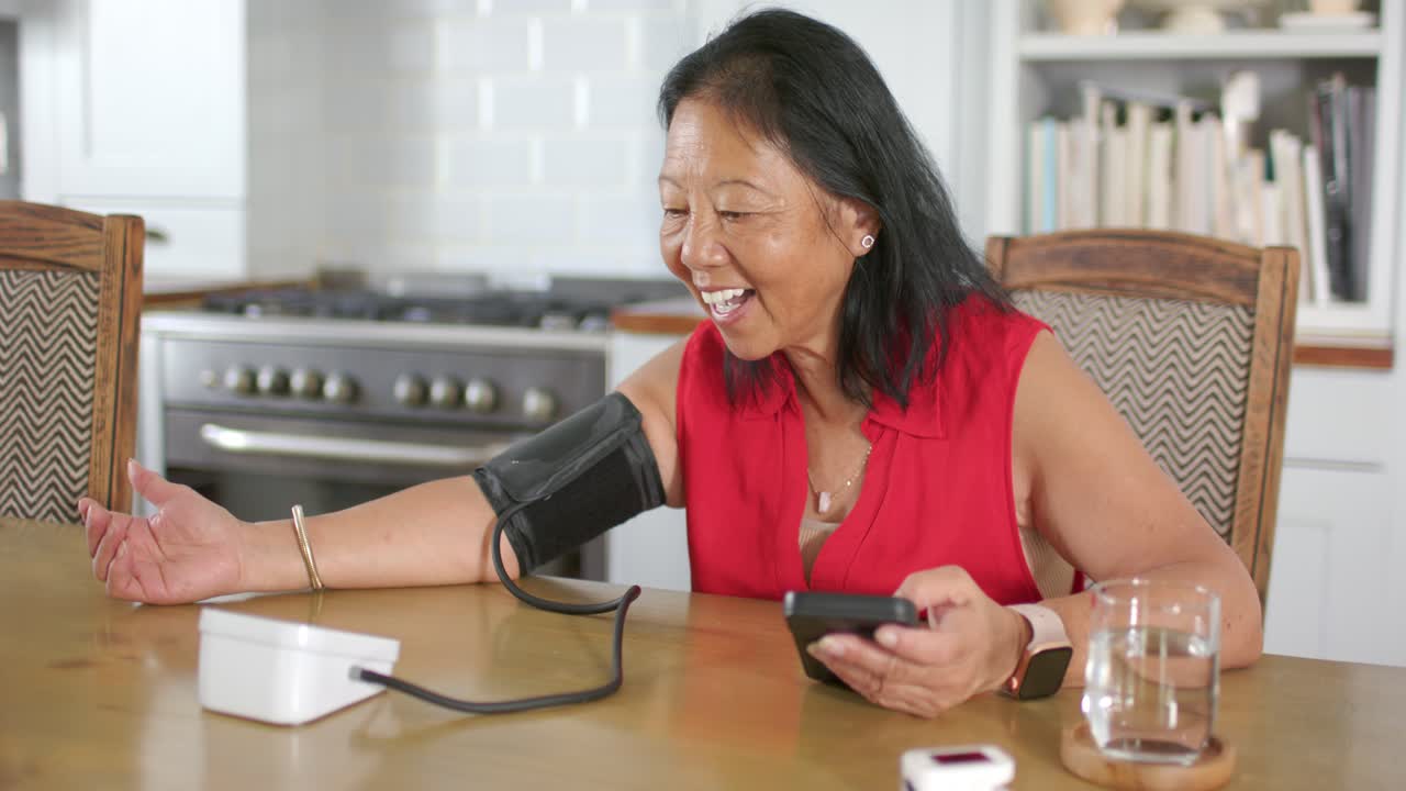 Anziana donna asiatica con una camicetta rossa seduta al tavolo della cucina, che usa il telefono e monitora il bracciale per la pressione sanguigna.