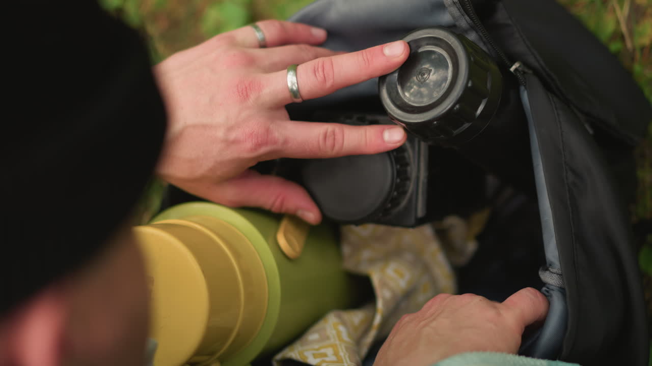 caucasian photographer retrieving camera from backpack, closeup hands removing lens cap and adjusting strap, intimate fieldwork scene with beanie and flannel, focused posture during nature excursion