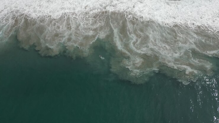 High aerial looks down onto tiny lone surfer in muddy shore waves