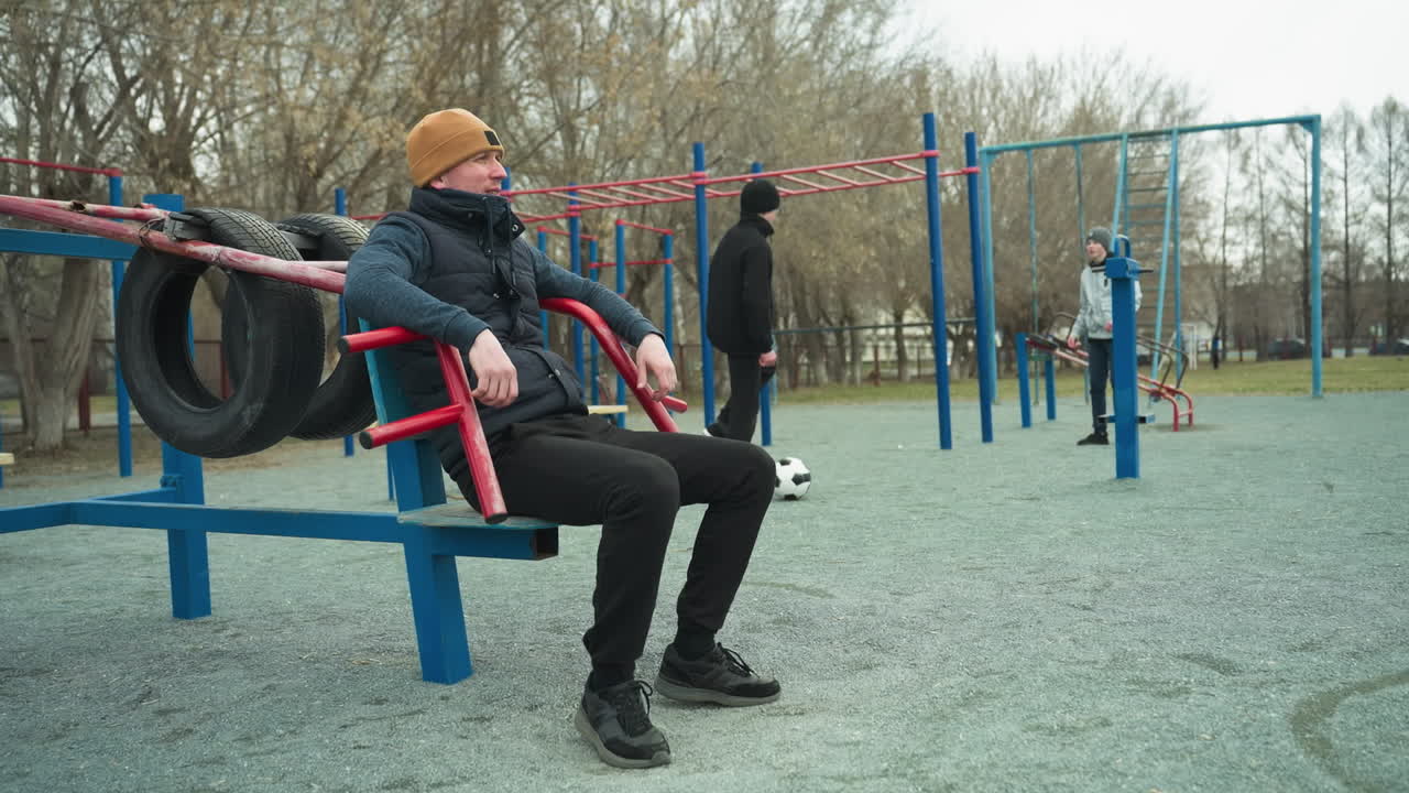 A coach is seated on workout equipment, casually shaking his leg, in the background, two boys are happily playing football and having a conversation, as one adjust backward