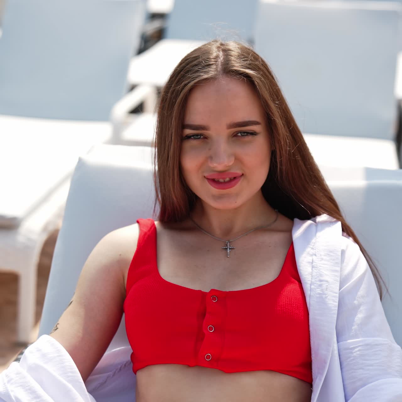 Giovane e sexy ragazza in costume rosso e camicia bianca che si gode il sole. La ragazza è sdraiata su una sedia a sdraio bianca e guarda nella fotocamera