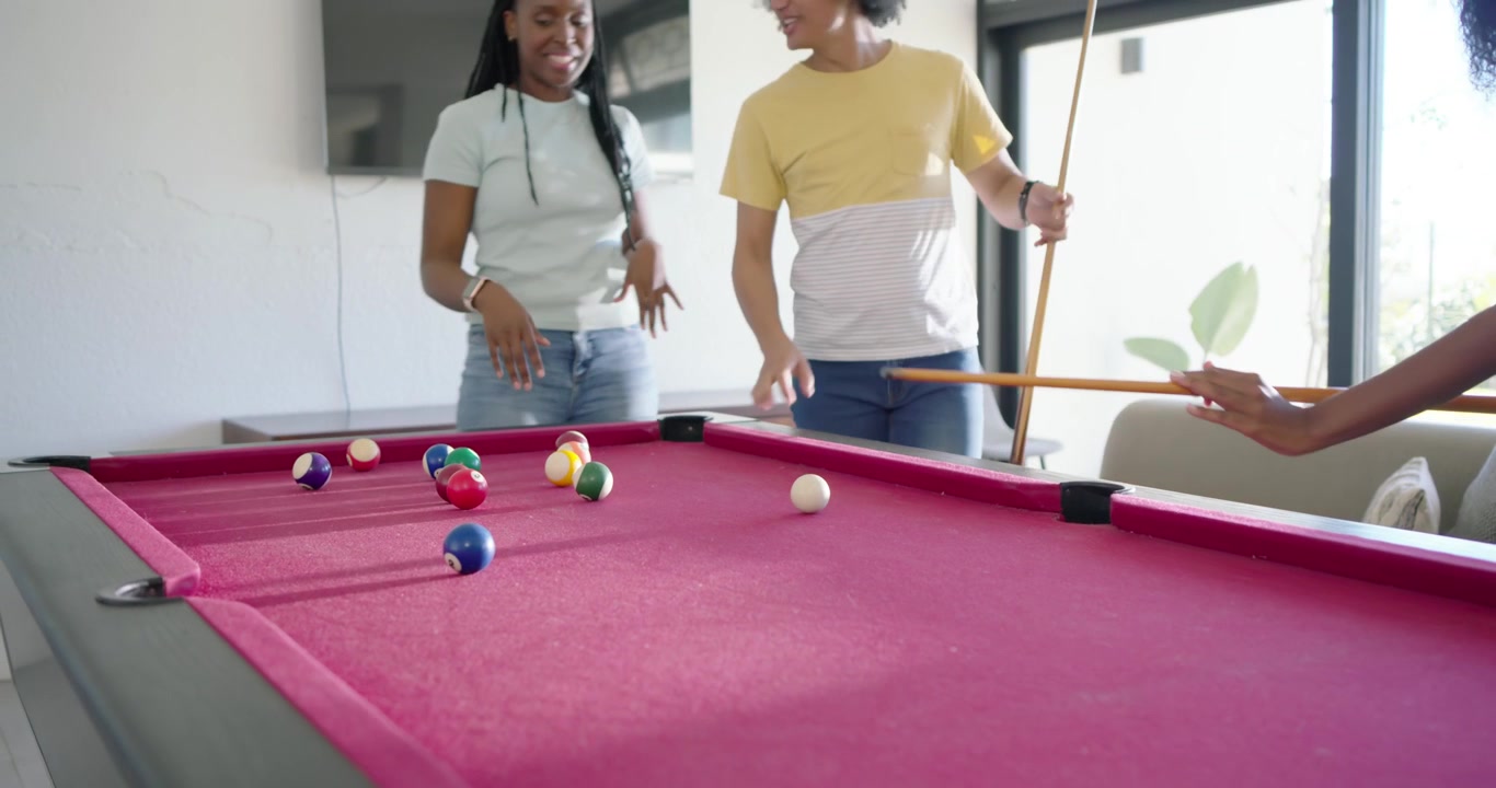 Diverse friends gathering at rec room pink-felt table after break, woman in pink top executing shot
