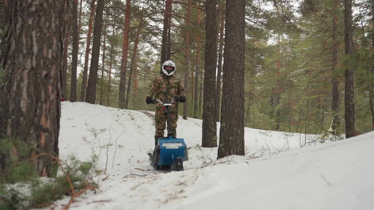 ATV ribaltato nella neve vicino agli alberi, pilota mimetizzato ispeziona la macchina blu danneggiata alla base del pendio, primo piano del cingolo e del sottocarro rivela componenti piegati, tentativi di recupero e riparazione, pineta silenziosa