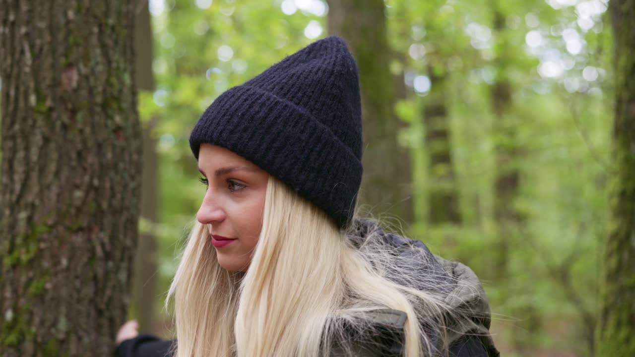 A young woman with long blonde hair wearing a black beanie and outdoor jacket walks through a green forest. She gently touches trees covered with moss while looking around with a peaceful expression
