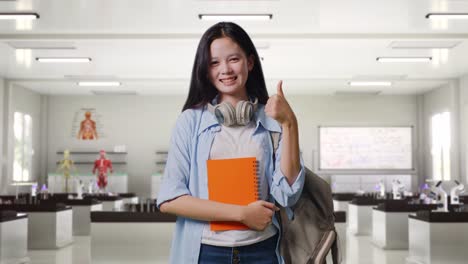estudiante adolescente asiática con una mochila y algunos libros sonriendo y mostrando los pulgares hacia arriba gesto a la cámara mientras está de pie en el laboratorio de ciencias