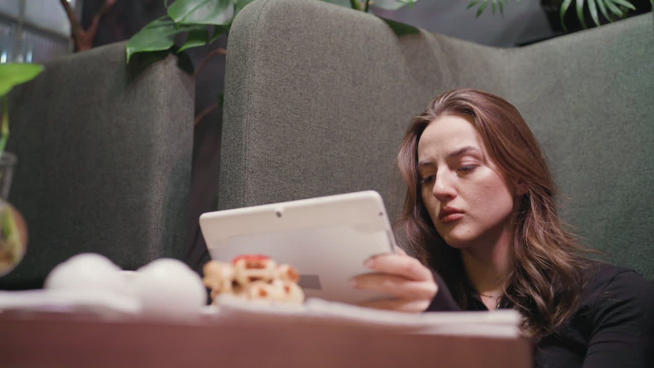 Focused lady operates notepad with concentrated expression while seated in cushioned lounge, surrounded by indoor plants and decoration, giving calm, tech-savvy, modern ambiance with dessert on table