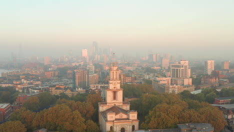 Toma-Aérea-De-Cerca-Del-Limehouse-De-La-Iglesia-De-St-Annes-Al-Amanecer-Con-El-Horizonte-Del-Centro-De-Londres-Al-Fondo