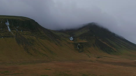 drone view of overcast skies, multiple mountains, brush covered ground, valleys and small patches of snow in iceland