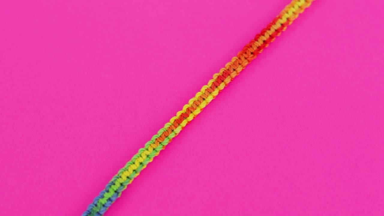 fotografía en primer plano de una colorida y vibrante pulsera de macrame roscada con patrón de color arco iris, descansando sobre un fondo rosa neón