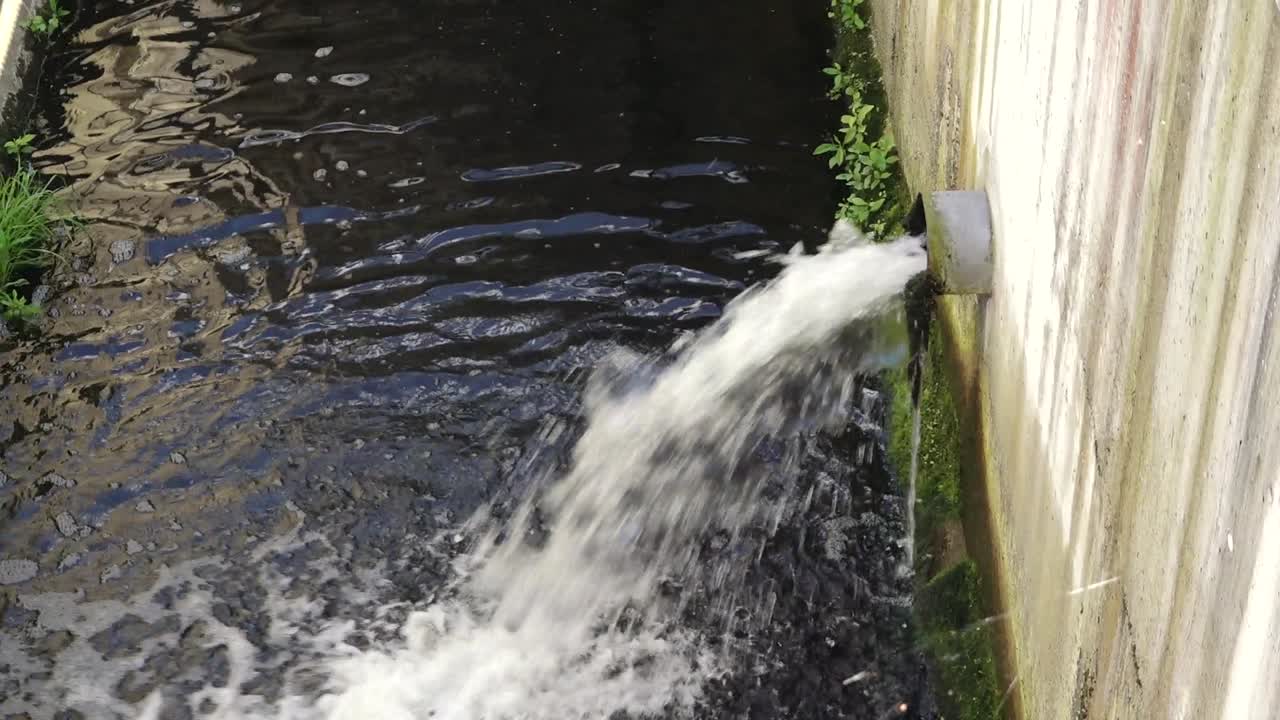 Water flowing strongly from a drainage pipe into a canal, creating ripples and foam on the dark surface of the water