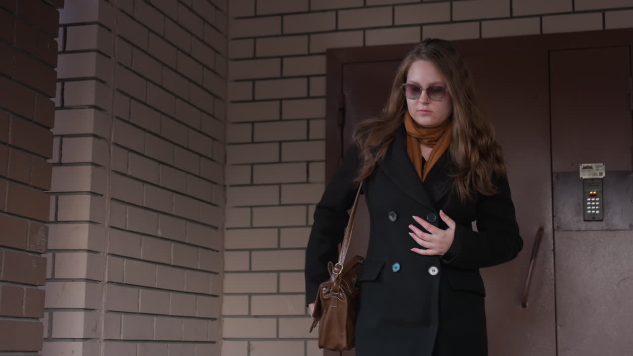 어두운 코트와 선글라스를 낀 세련된 여성이 문에서 나와 갈색 가죽 가방에 손을 넣고, 스카프가 흔들리는 모습이에요. 벽돌 벽 배경에 자연스러운 도시의 한순간, 목적 있는 움직임, 일상적인 라이프스타일 장면이 담겨 있어요.