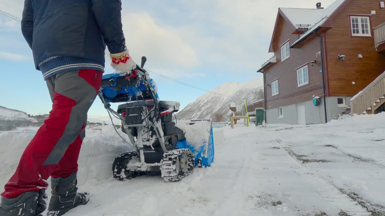 Mand rydder sne med en sneblæser i indkørslen, efter at sneploven flyttede al sneen tilbage på privat grund, med smukke bjerge i baggrunden.