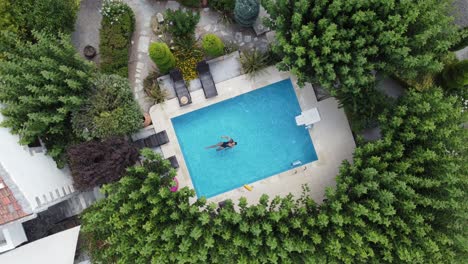 young woman enjoying summer in pool - floats on back - drone hovers over pool in bird's eye view