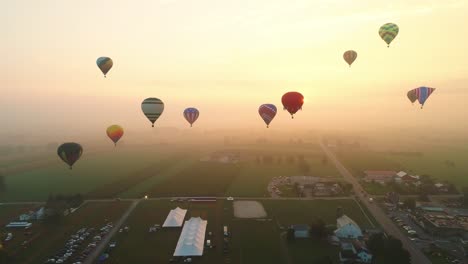 an aerial view of multiple hot air balloons rising into the early mist during a festival with crowds watching, on a summer day