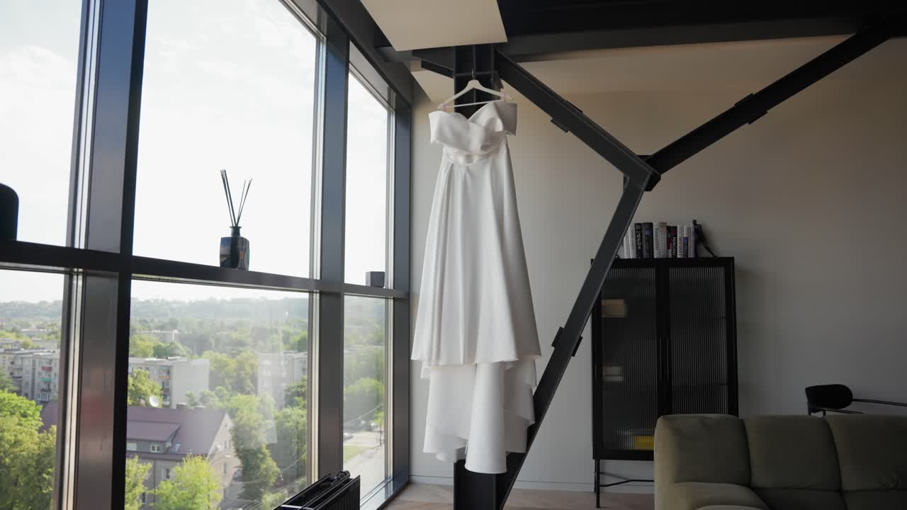 White wedding dress hanging in a modern apartment interior by large glass windows, with scenic daylight and city view in the background, capturing elegance and bridal preparation