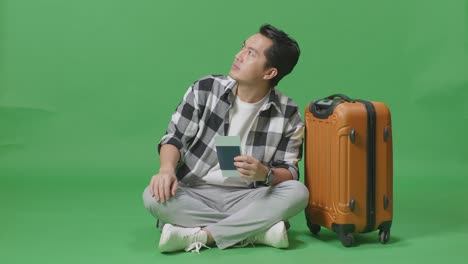 full body of asian male traveler with luggage looking at the passport and looking around while sitting in the green screen background studio