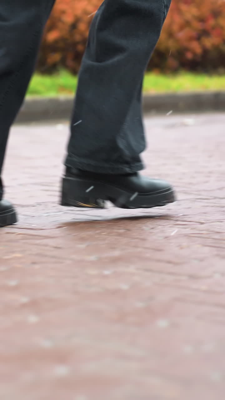 Close-up shot of human legs running during light snowfall, wearing black jeans and black boots. Background features autumn trees with leaves and wintery atmosphere, creating a serene feel