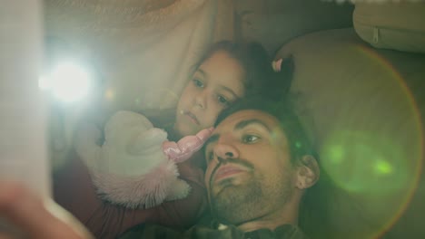 Close-up-shot-of-a-brunette-man-in-a-green-checkered-shirt-reading-a-fairy-tale-to-his-daughter-under-a-makeshift-awning-at-home-near-a-gray-sofa.-A-little-brunette-girl-lies-under-a-makeshift-awning-at-home-and-listens-to-her-father’s-stories-with-a-unicorn-toy-in-her-hands