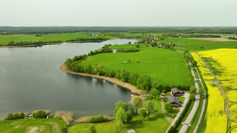 Aerial-view-of-a-lake-surrounded-by-green-fields,-small-houses,-and-a-rapeseed-field,-under-a-clear-sky