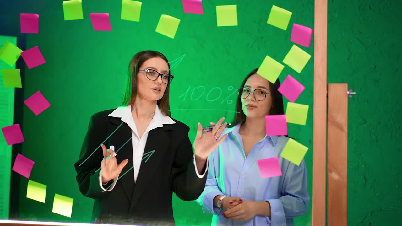 Attractive women working on ideas in the office. Ladies use glass board, write on it and look at stickers. Brainstorming concept.