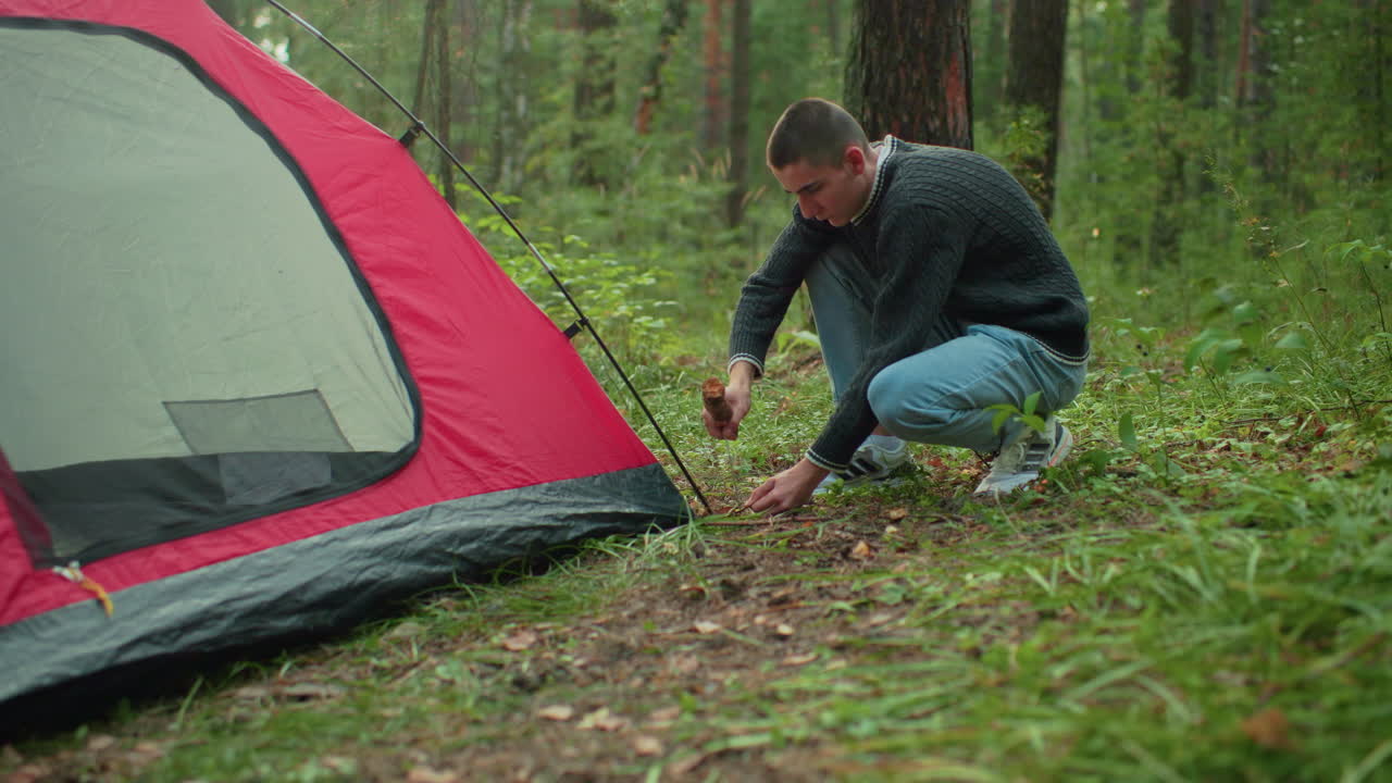 student squats beside pitched red and gray tent in forest holding thick wood while hammering peg into grassy ground with focused expression surrounded by trees and lush greenery