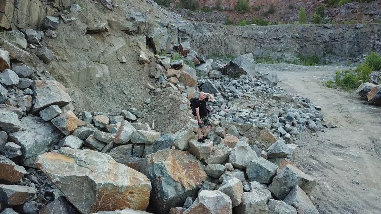 A tourist in shorts and a backpack is standing on a huge stone in the middle of a quarry and looking into the distance to the surrounding landscape. Camera motion up. Aerial view.